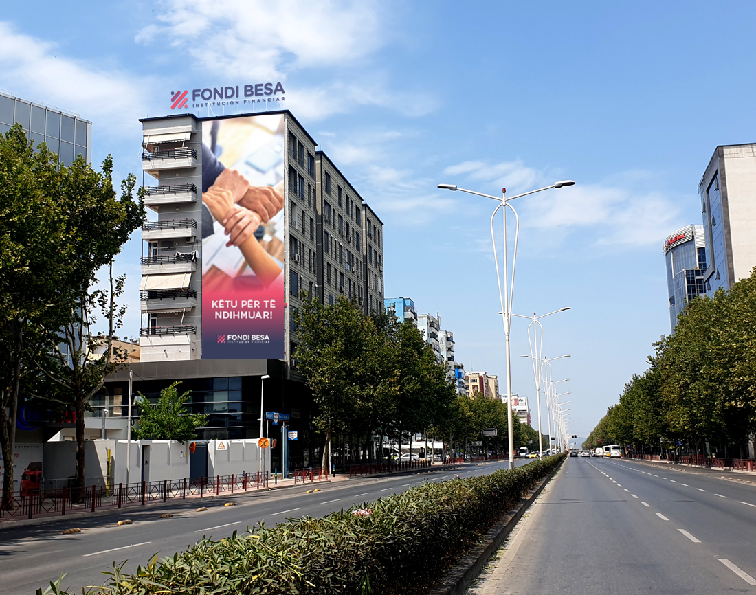 Street-level view of the Fondi BESA headquarters in Tirana, Albania, featuring the institution's logo and a large banner on the building façade, with a wide tree-lined boulevard in the foreground.